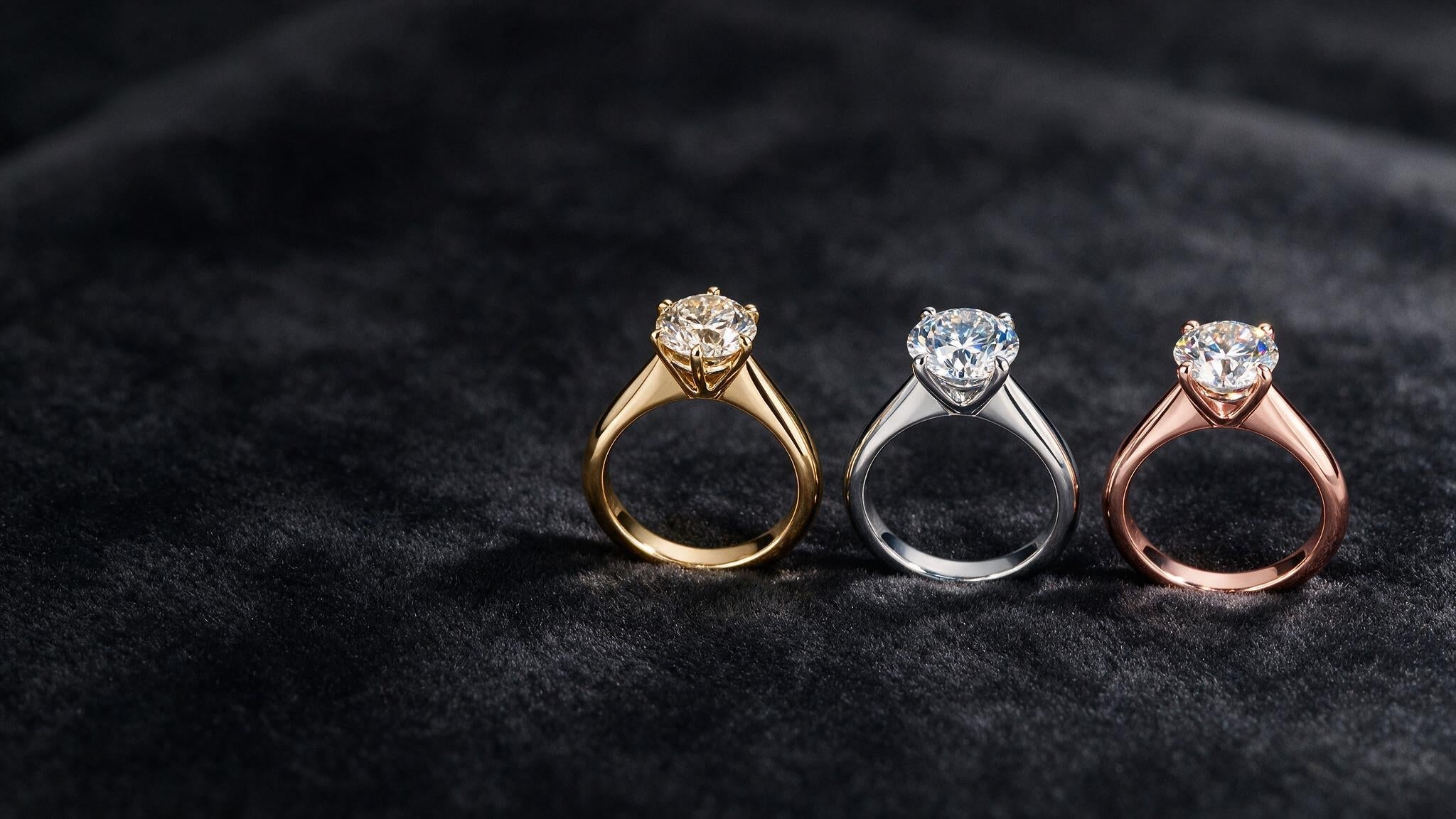 Three diamond solitaire rings in yellow, white, and rose gold side by side on dark velvet